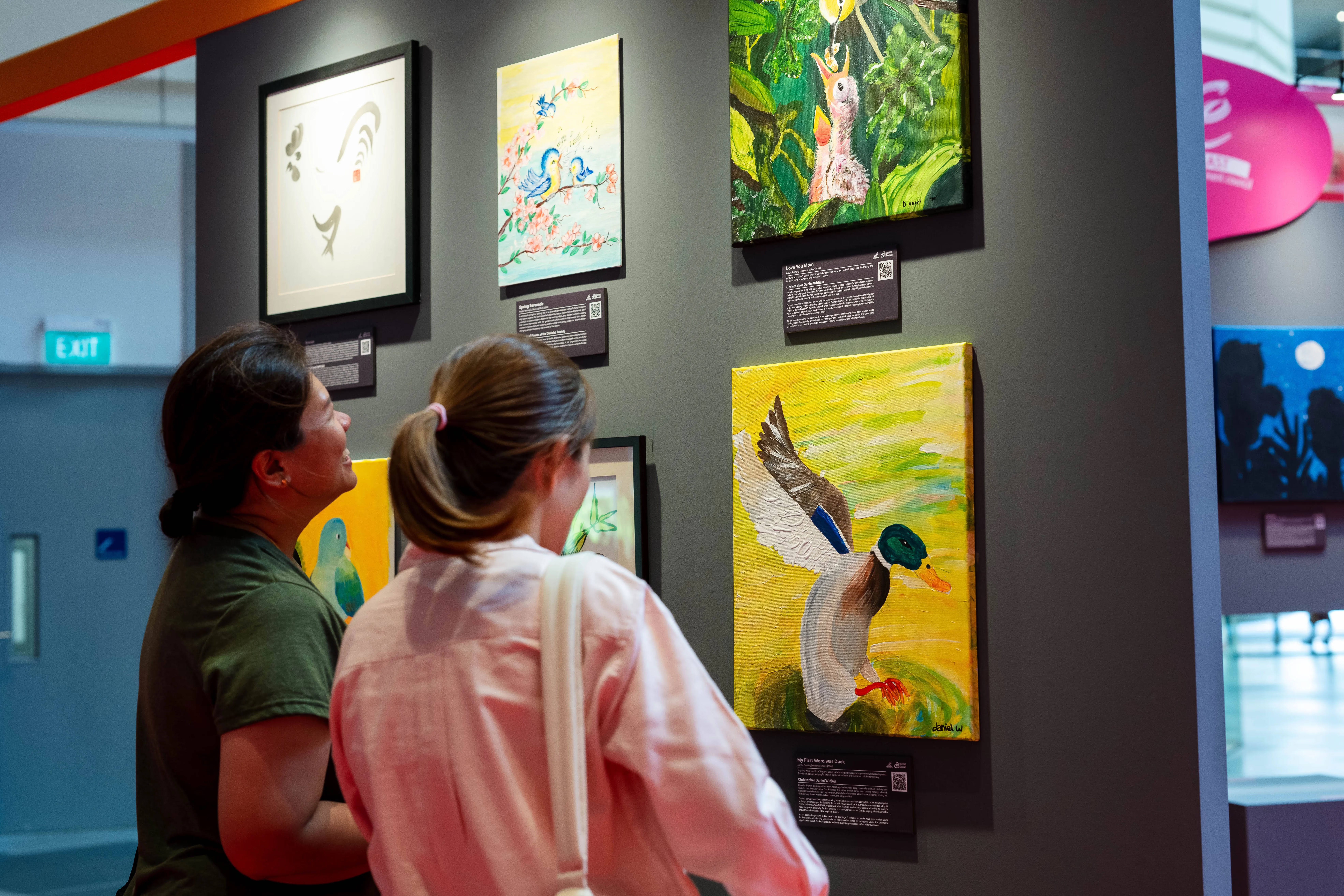 Two individuals are viewing various paintings, including birds, displayed on a dark wall in an art exhibition.